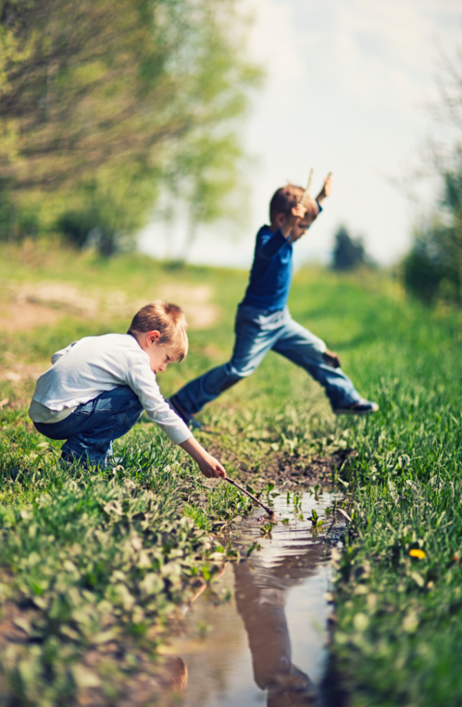 Two young children playing outside in the summer. One boy plays with a stick in some water while another leaps over the stream.